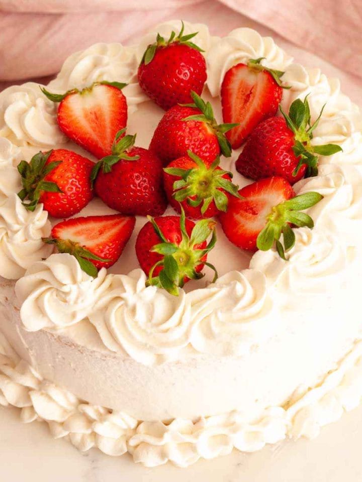 A strawberry whipped cream cake on a marble surface next to a pink towel and topped with fresh strawberries.