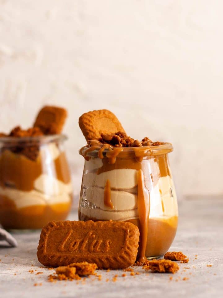 A close up of two Biscoff cheesecake cups in small tulip jars on a gray surface next to crushed Lotus Biscoff cookies.