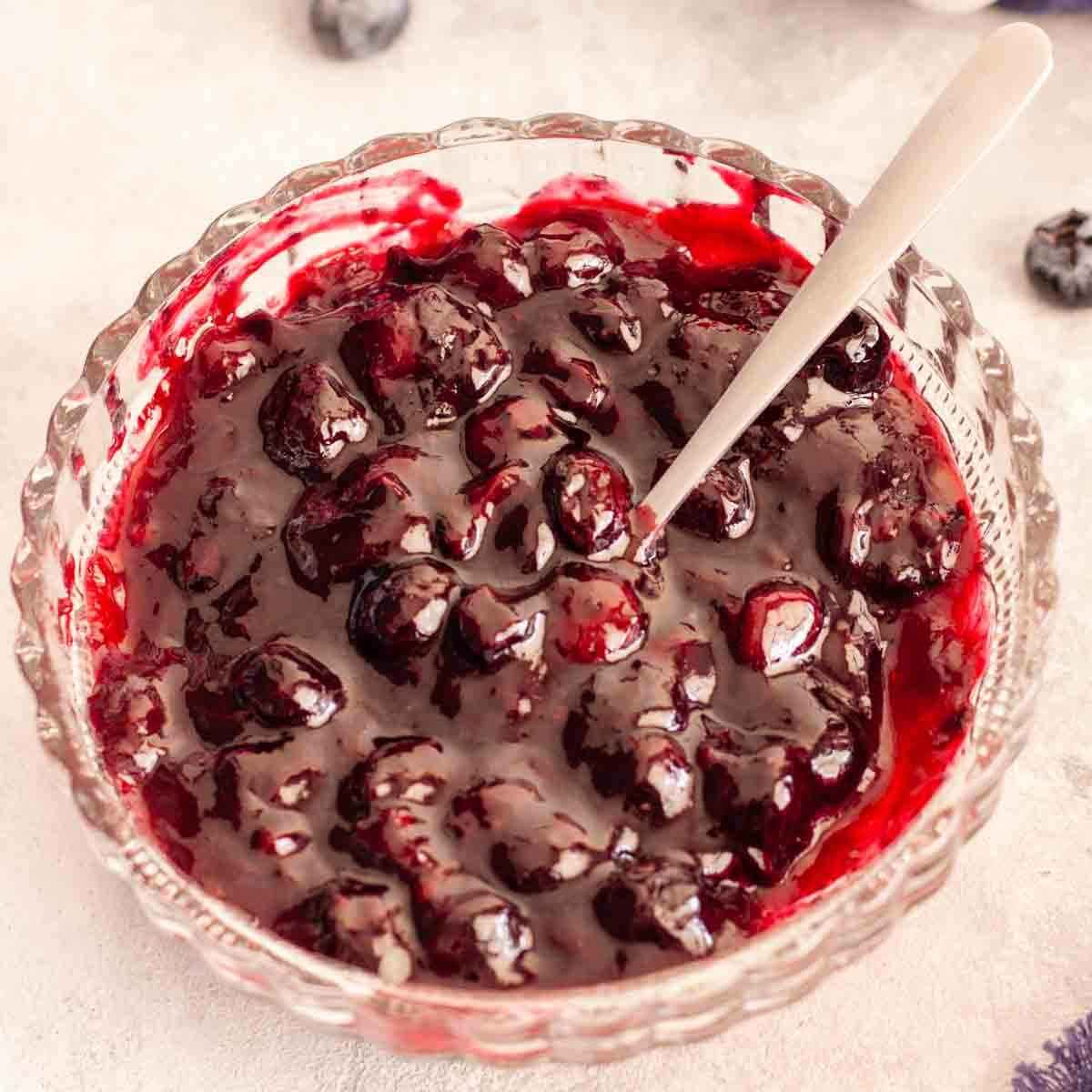 Blueberry cake filling in a glass bowl with a spoon on a gray surface next to blueberries.