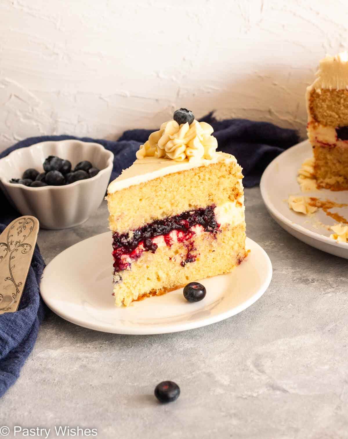 A slice of vanilla cake filled with blueberry filling next to a bowl of blueberries and cut layer cake.