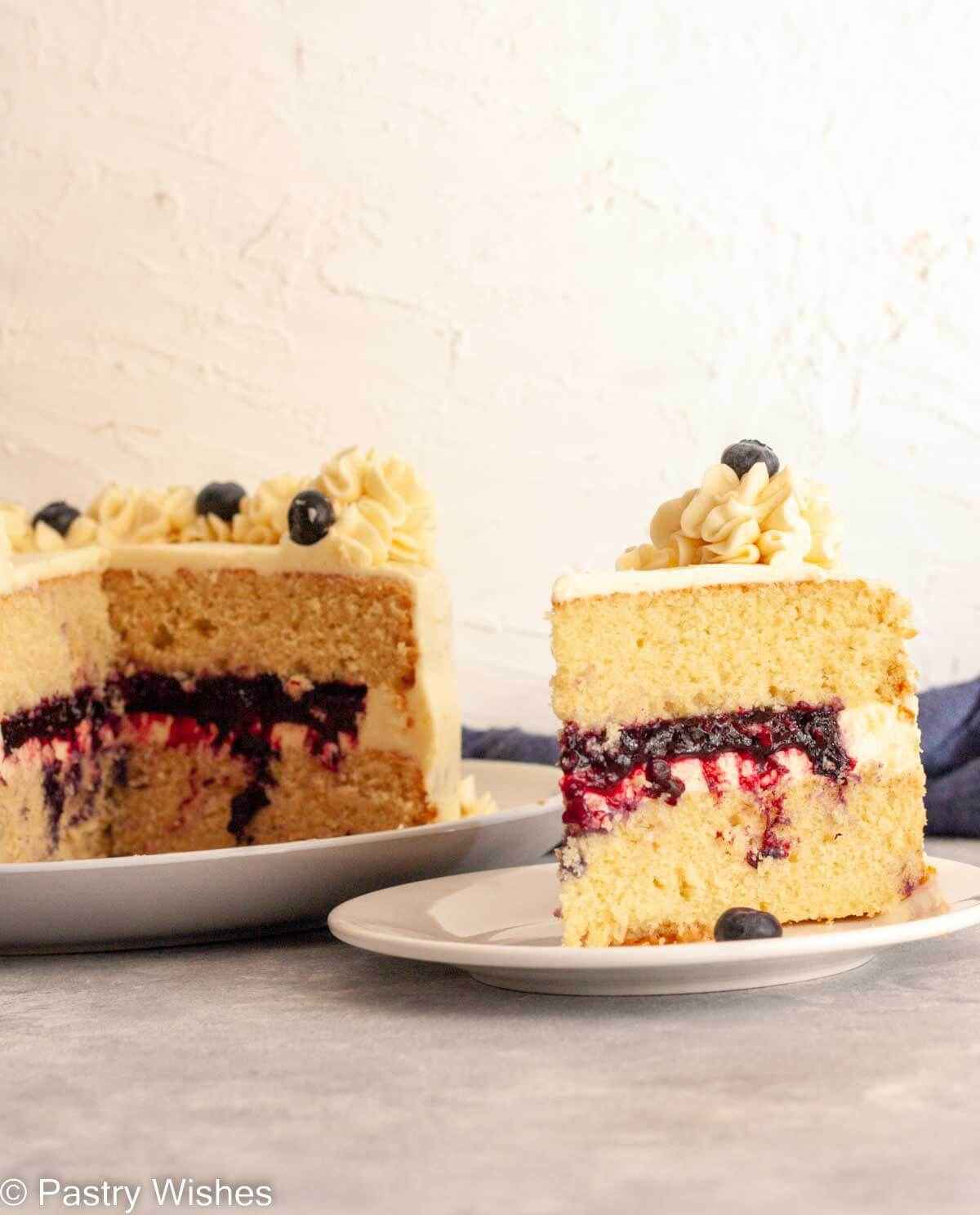 A slice of vanilla cake with blueberry filling on a white plate next to a layer cake filled with blueberry filling.