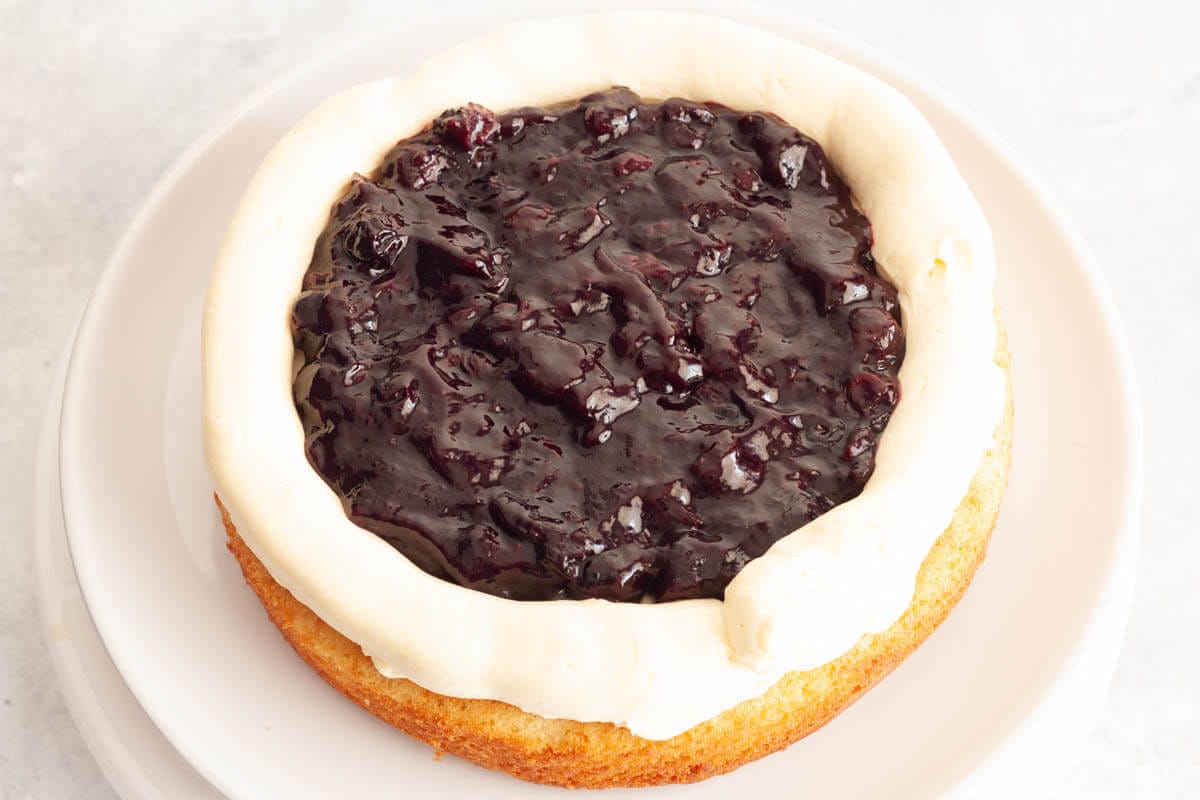 A vanilla cake layer on a white plate topped with vanilla buttercream frosting and a border of frosting around the edge and filled with blueberry cake filling.