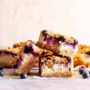 A closeup of three blueberry cheesecake crumble bars on a gray and white surface next to fresh blueberries.