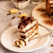 A closeup of a slice of chocolate tiramisu cake with a fork on a white plate next to ladyfingers, coffee beans and cocoa powder dusted on serving plate with cake.