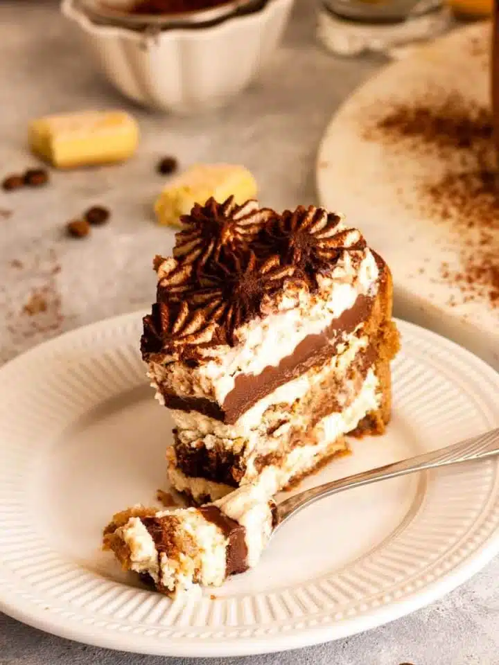 A closeup of a slice of chocolate tiramisu cake with a fork on a white plate next to ladyfingers, coffee beans and cocoa powder dusted on serving plate with cake.