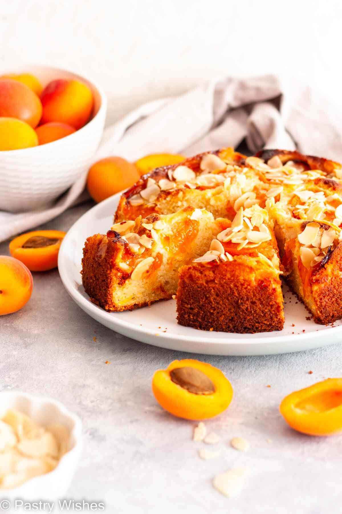 Sliced apricot almond cake on a white plate next to a bowl of fresh apricots and sliced almonds.