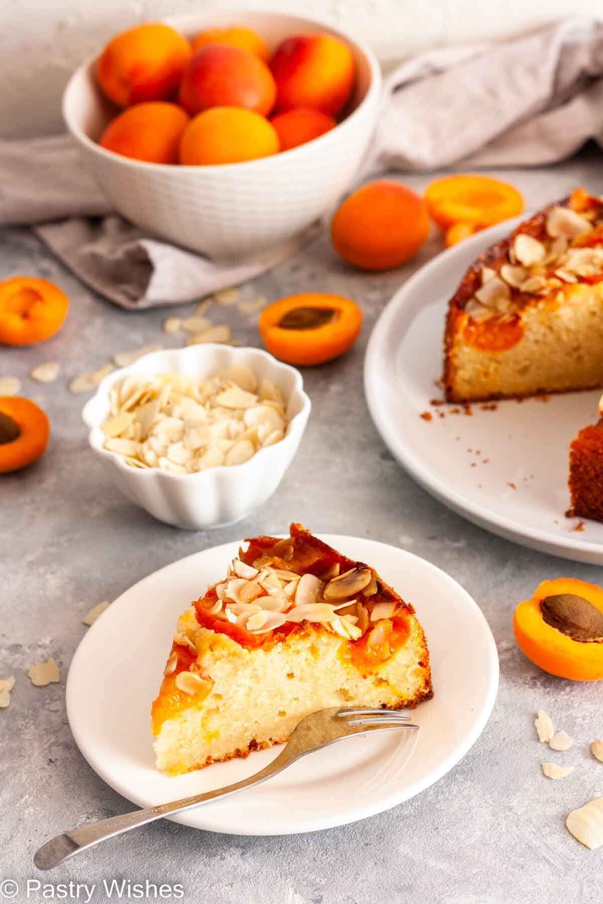 A slice of apricot almond cake on a white plate with a fork next to a bowl of fresh apricots, a gray kitchen towel and a bowl of sliced almonds.