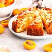 A close-up of sliced apricot almond cake on a white plate next to a bowl of fresh apricots and sliced almonds.