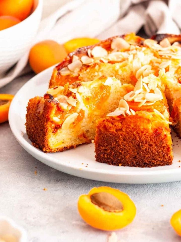 A close-up of sliced apricot almond cake on a white plate next to a bowl of fresh apricots and sliced almonds.