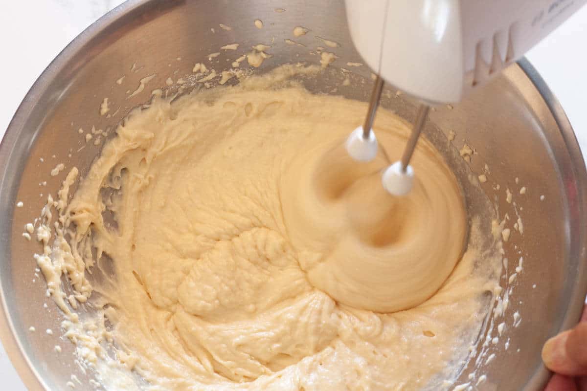 Beating together wet ingredients and dry ingredients with a hand mixer in a metal mixing bowl.