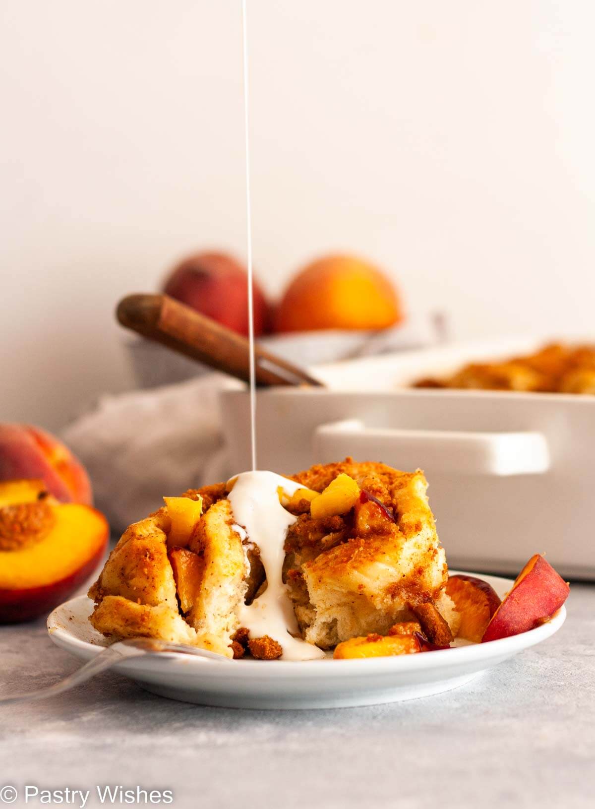 Pouring cream over a peach cinnamon roll on a white plate with fresh peach slices.