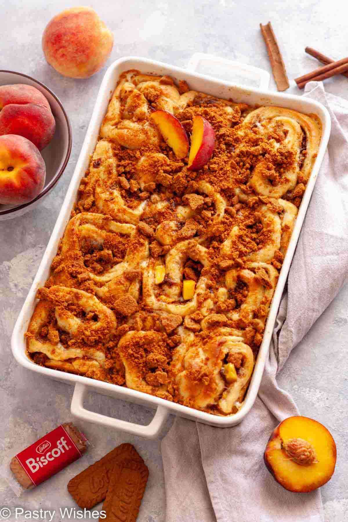 Peach cinnamon rolls topped with crushed Biscoff cookies in a white baking dish next to a gray kitchen towel, a bowl of peaches, and cinnamon sticks.