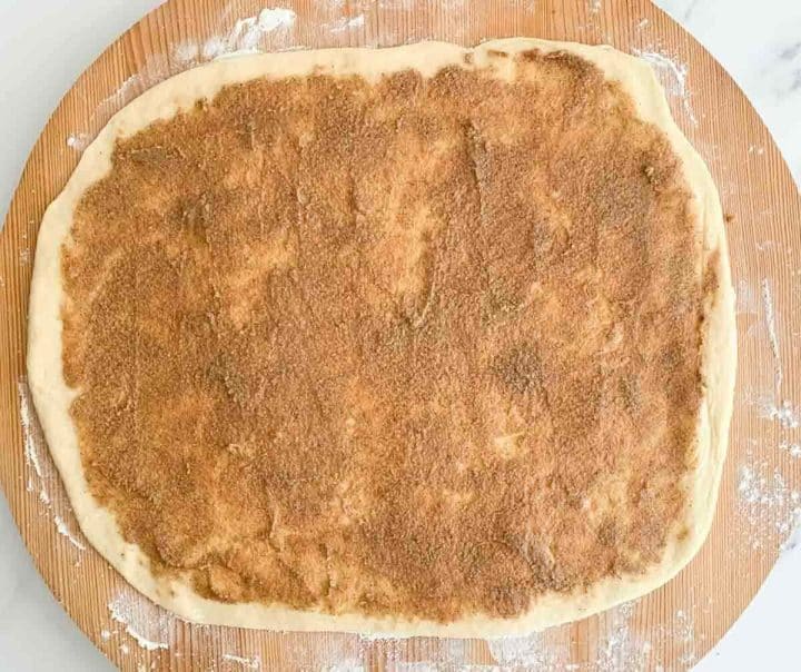 Cinnamon sugar butter spread over dough on a floured wooden surface.