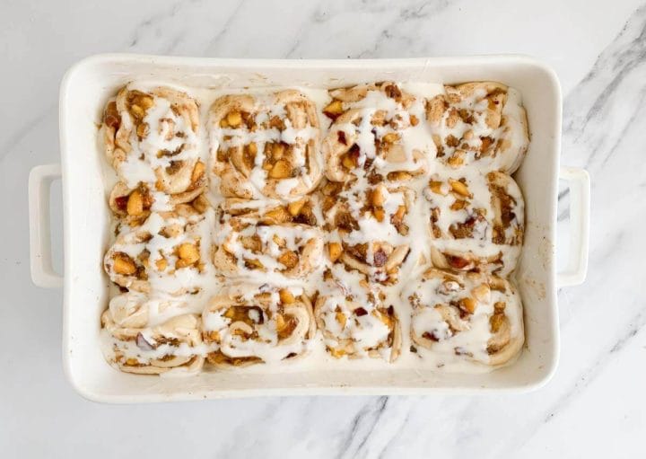 Heavy cream poured over twelve peach cinnamon rolls in a white baking dish.