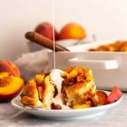 Pouring cream over a peach cinnamon roll on a white plate with fresh peach slices.