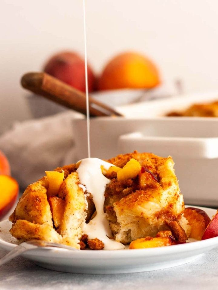 Pouring cream over a peach cinnamon roll on a white plate with fresh peach slices.