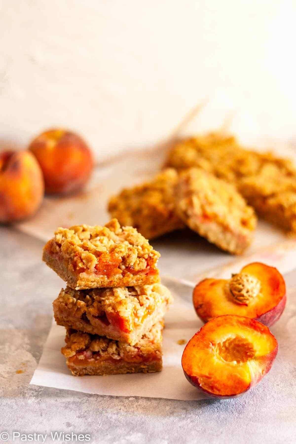 A stack of peach crumble bars next to fresh peaches with more peach crumble bars in the background.