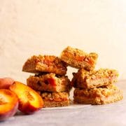 A close of of two stacks of peach crumble bars next to sliced peaches.