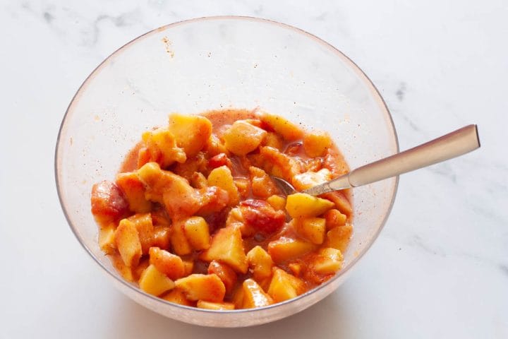 Chopped peaches in a glass bowl with a spoon mixed with sugar, cornstarch, lemon juice and ground cinnamon.