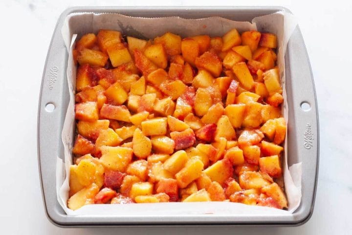 Peach filling over a baked crust in a square pan lined with parchment paper.