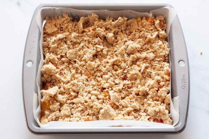 Crumble topping over peach filling in a square pan lined with parchment paper.