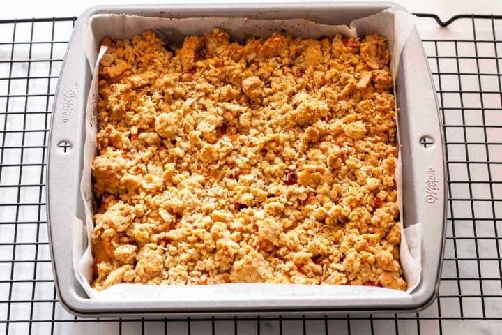 A baked peach crumble dessert in a square pan lined with parchment paper on a wire rack.