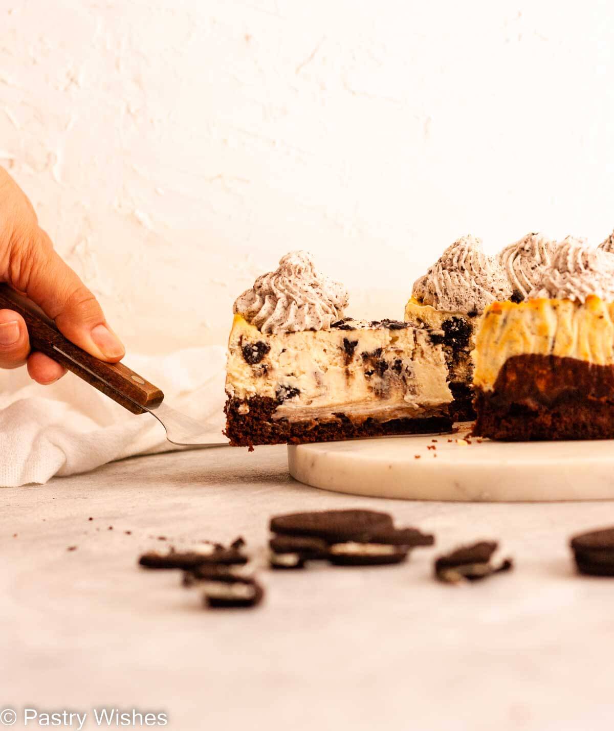 A slice of Oreo brownie cheesecake on a cake server topped with Oreo whipped cream, nest to Oreo cookies.