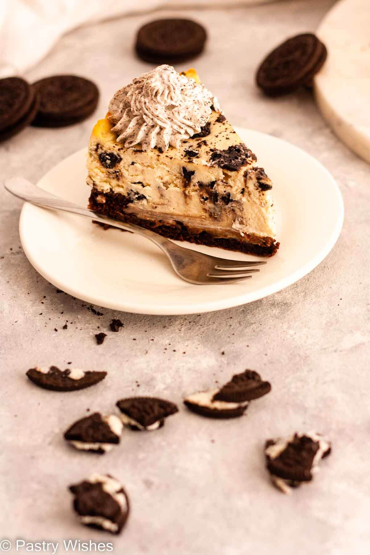 A slice of Oreo brownie cheesecake topped with Oreo whipped cream on a white plate with a fork next to Oreo cookies.