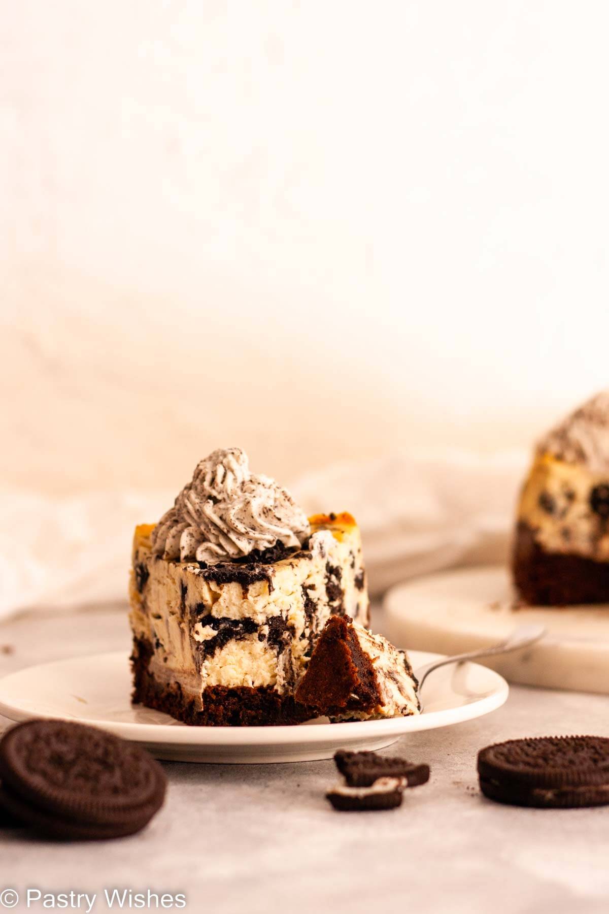A slice of Oreo brownie cheesecake topped with Oreo whipped cream on a white plate with a fork next to Oreo cookies.