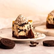 A close up of a slice of Oreo brownie cheesecake topped with Oreo whipped cream on a white plate with a fork next to Oreo cookies.