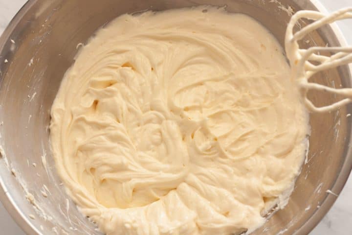 Beating eggs, Greek yogurt, and vanilla with the cream cheese mixture using a hand mixer in a metal bowl.