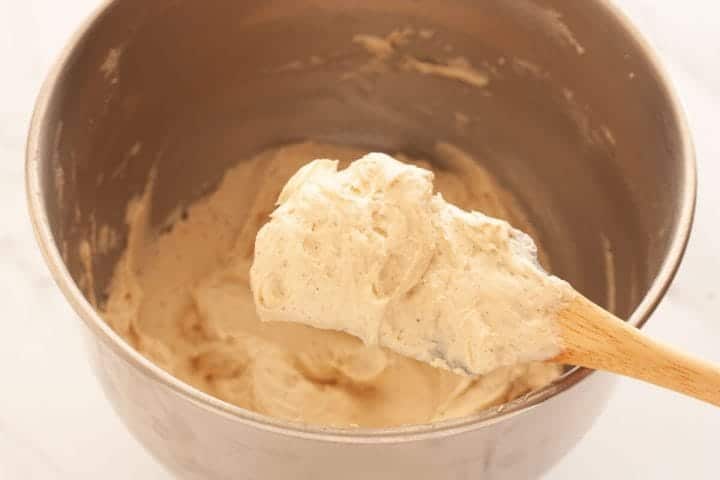 A close up of a rubber spatula with some cinnamon buttercream over a metal bowl full of buttercream.