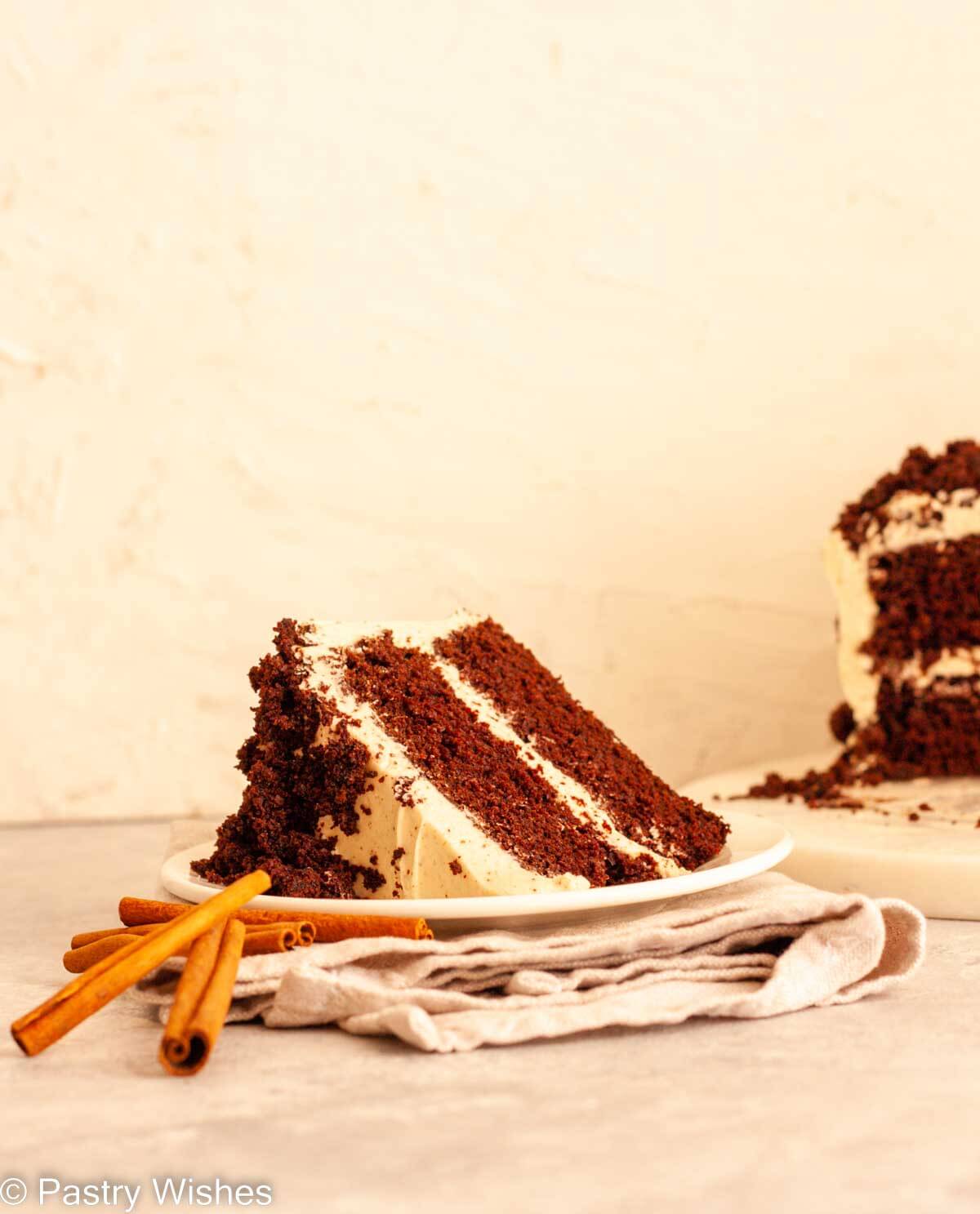 A slice of spiced chocolate cake on a white plate on a folded gray napkin next to cinnamon sticks.