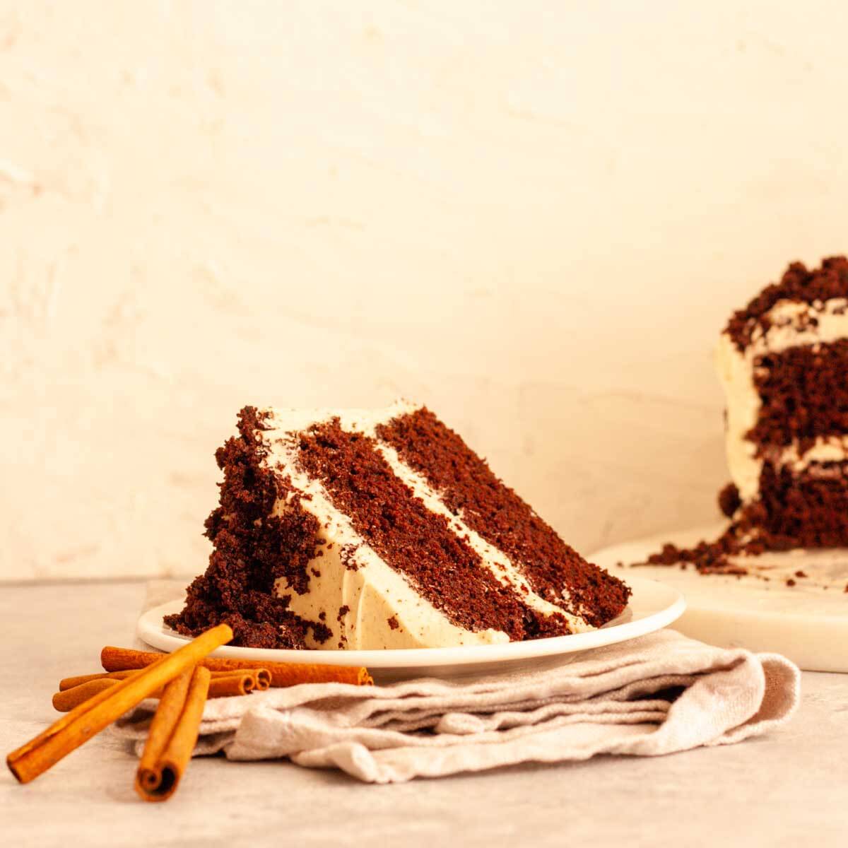 A slice of spiced chocolate cake on a white plate on a folded gray napkin next to cinnamon sticks.