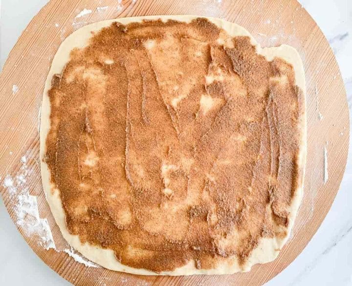 Cinnamon sugar filling over rolled out cinnamon roll dough on a wooden board.
