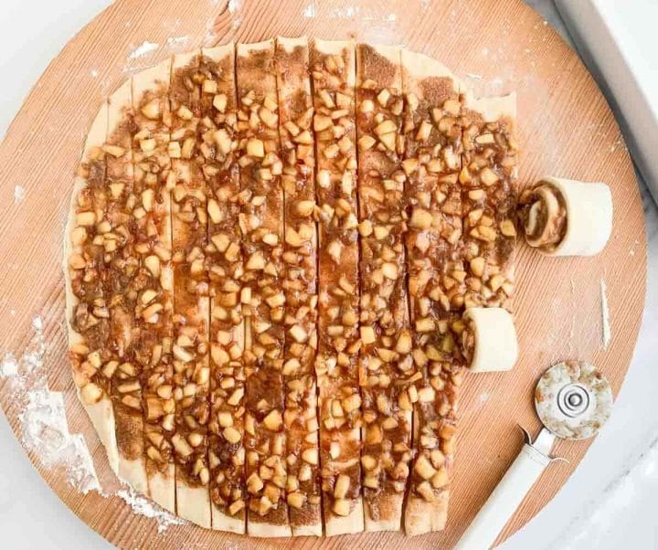 Apple pie cinnamon roll strips and a pizza cutter on a wooden board.