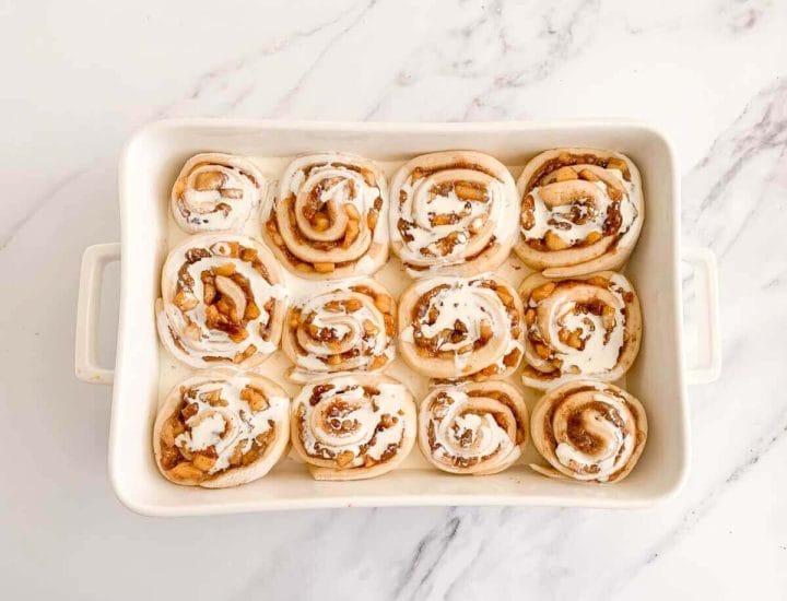 Warm heavy cream poured over risen apple cinnamon rolls in a baking dish before baking.