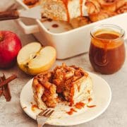 A closeup of an apple pie cinnamon roll on a white plate next to apples, cinnamon sticks, and a jar of caramel sauce with a pan of apple cinnamon rolls in the back.