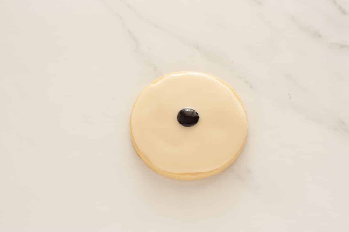 A small black dot of glaze in the center of a sugar cookie topped with white glaze on a sugar cookie.