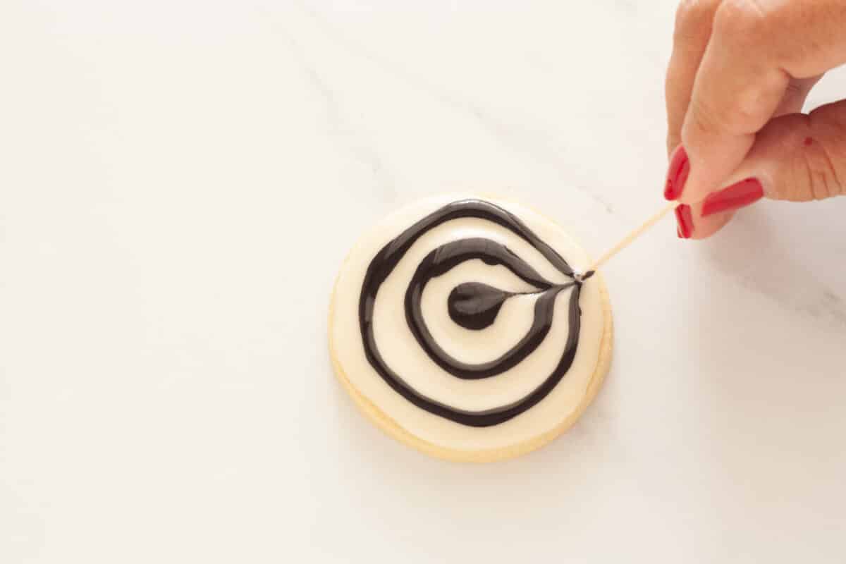 A hand holding a toothpick and dragging it from the center outward on a glazed cookie.