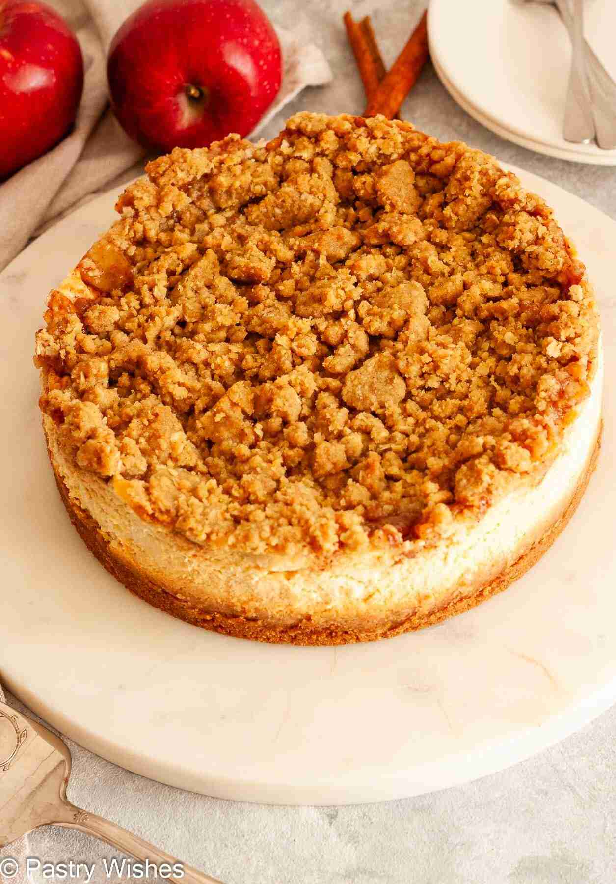 An overhead image of a whole apple crumble cheesecake next to apples, cinnamon sticks, plates and forks.