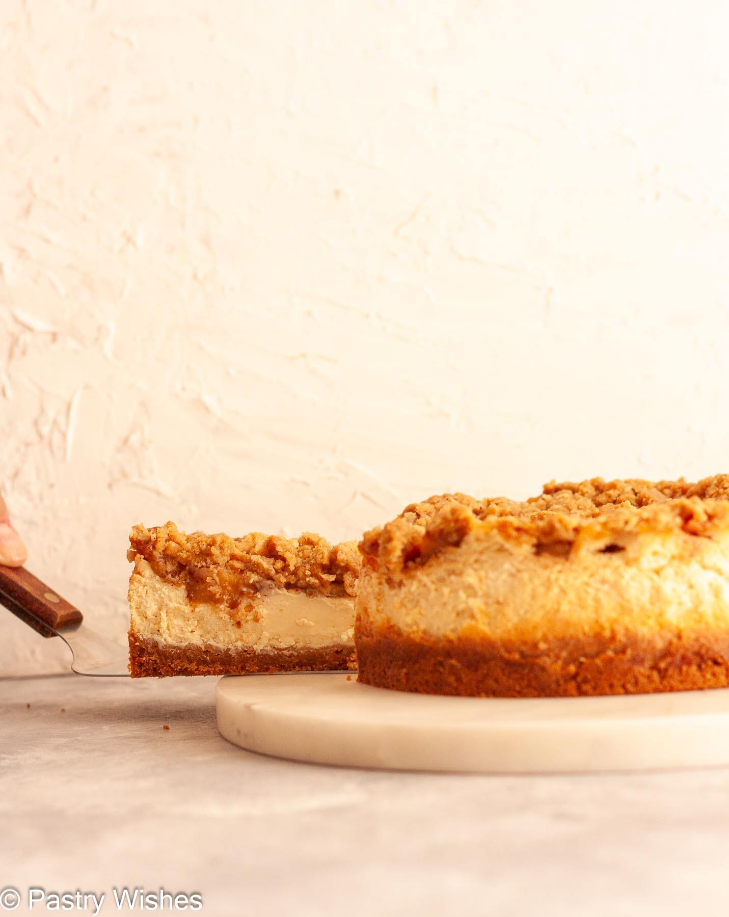 A hand holding a slice of apple crumble cheesecake on a cake slicer on a white marble platter.
