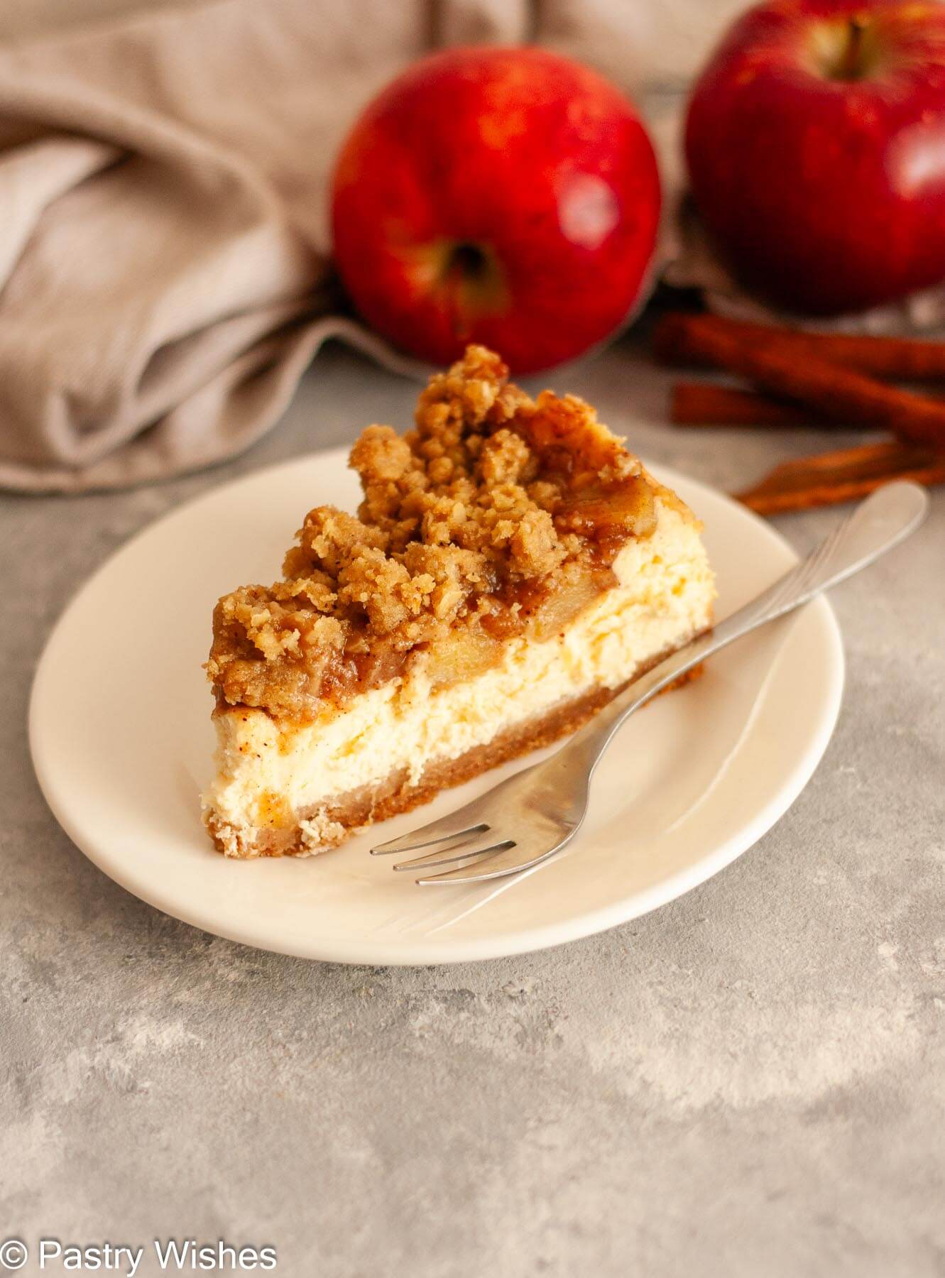 A slice of apple crumble cheesecake on a white plate with a fork next to apples, cinnamon sticks, and a gray kitchen towel on a gray and white surface.