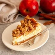A closeup of a slice of apple crumble cheesecake on a white plate with a fork next to apples, cinnamon sticks, and a gray kitchen towel.