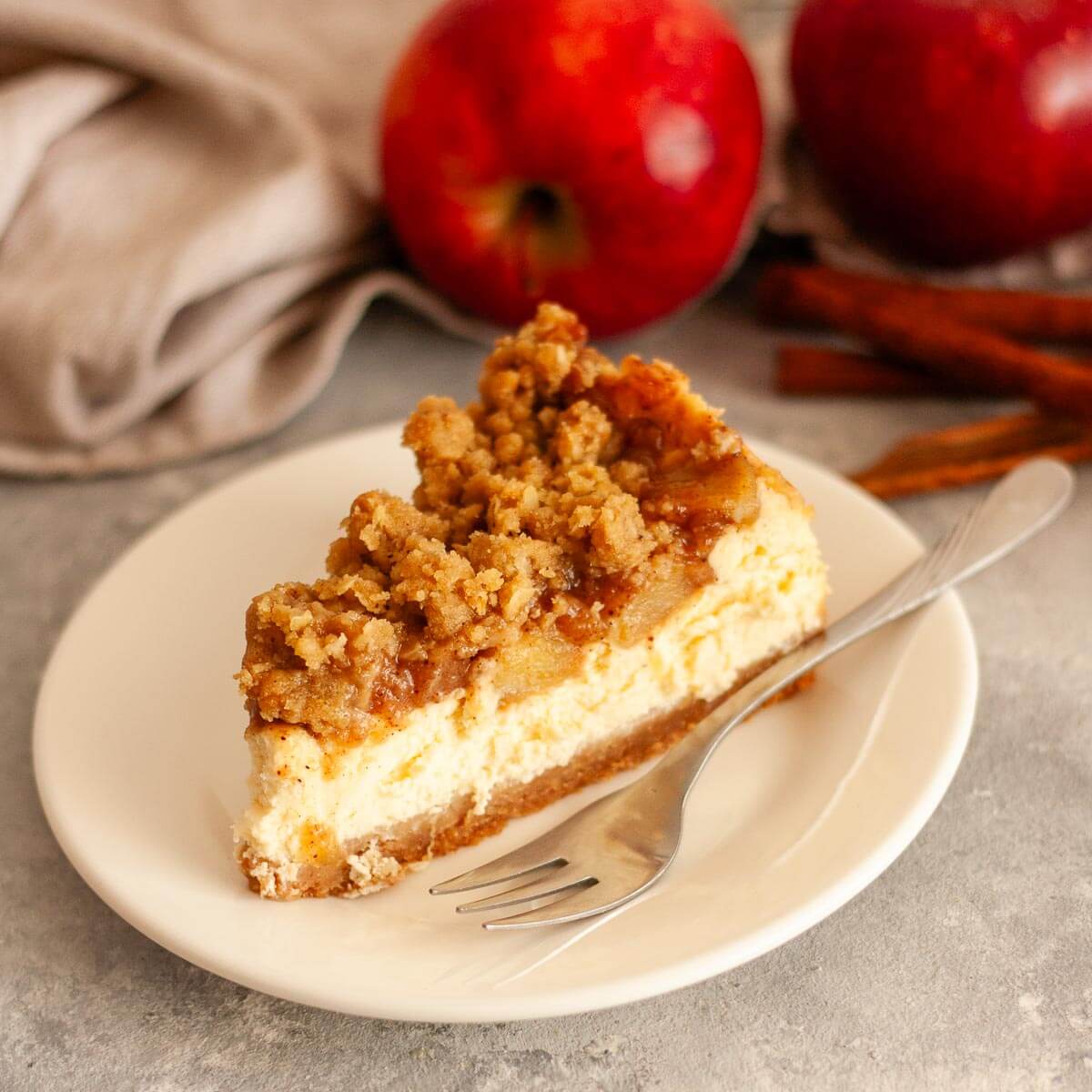 A closeup of a slice of apple crumble cheesecake on a white plate with a fork next to apples, cinnamon sticks, and a gray kitchen towel.