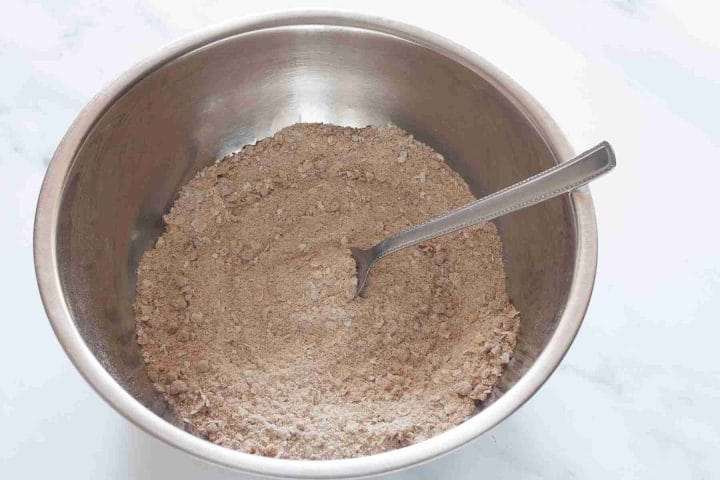 Flour brown sugar, rolled oats and spices in a metal bowl with a spoon.
