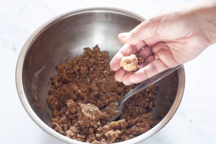 A hand holding a clump of crumble topping over a metal bowl.
