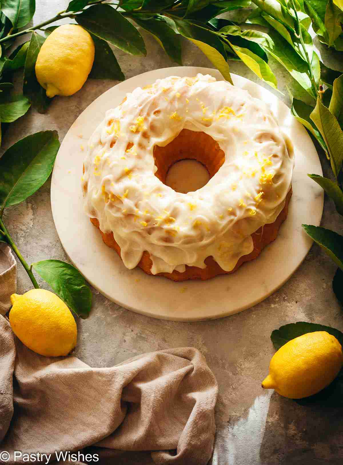Glazed lemon Bundt cake photographed from above with lemons, lemon tree branches, and a kitchen towel.