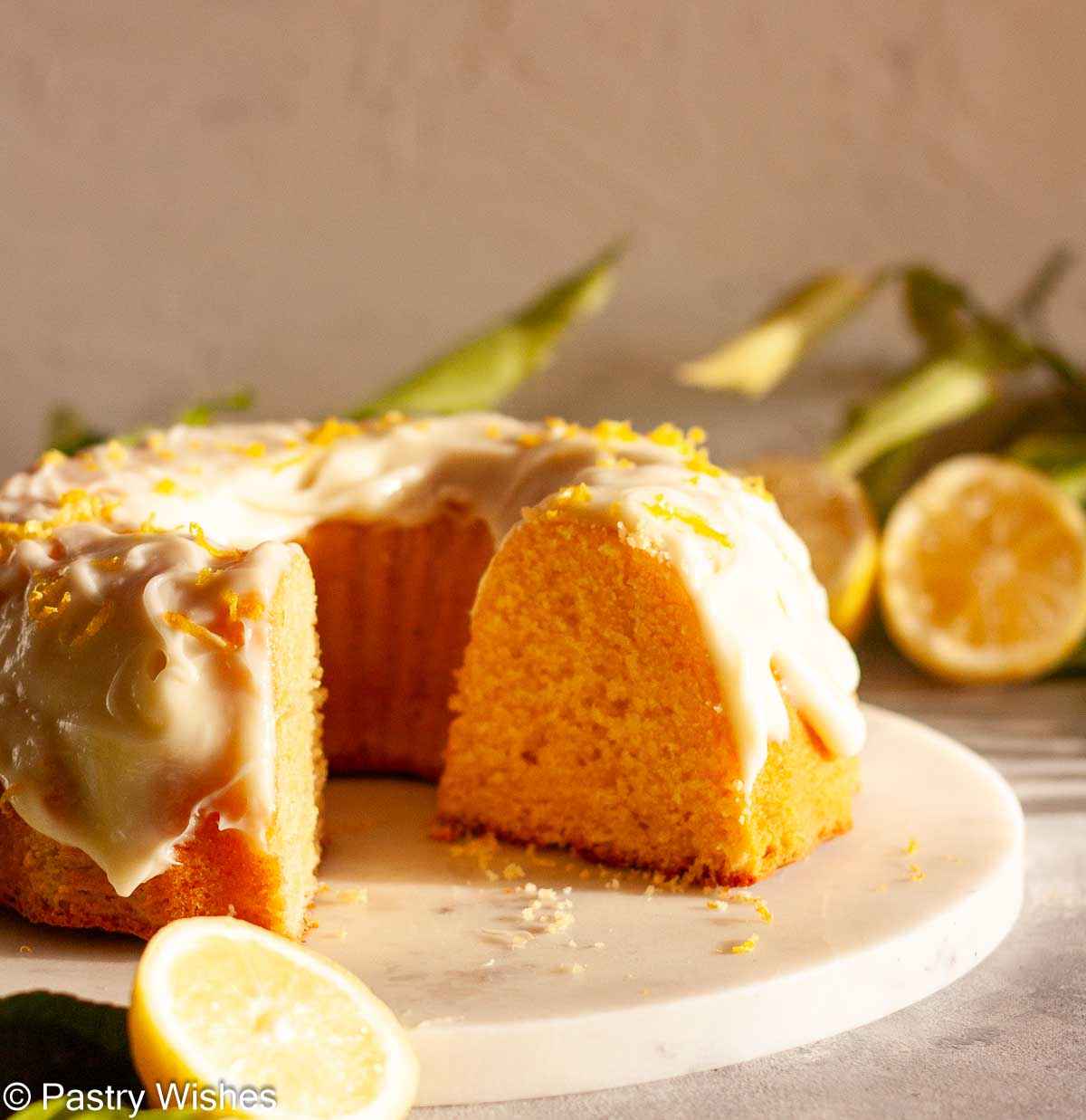 A close up of a cut lemon Bundt cake on a marble platter next to sliced lemons and lemon branches.