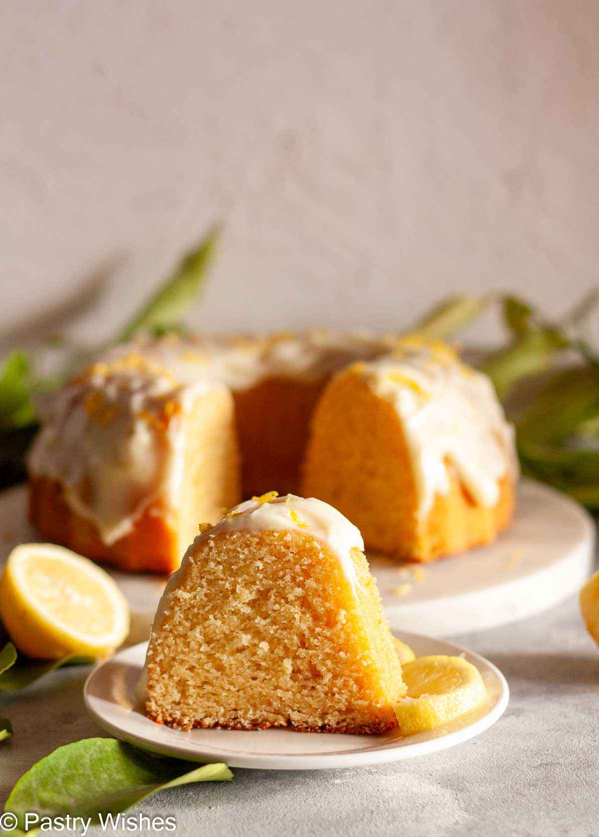 Wider view of a glazed lemon Bundt cake slice on a plate with the sliced cake in the background.
